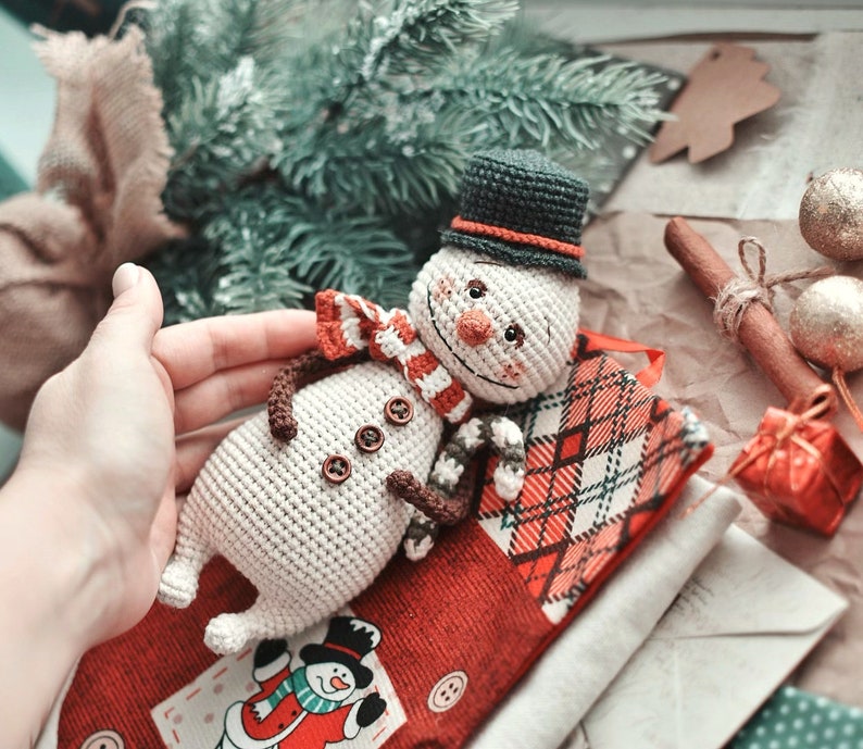 Puede incluir: Un adorno de mu&ntilde;eco de nieve de crochet con un sombrero negro, un cuerpo blanco y una bufanda a rayas rojas y blancas. El mu&ntilde;eco de nieve est&aacute; tumbado sobre una tela de cuadros rojos y blancos con un adorno de mu&ntilde;eco de nieve m&aacute;s peque&ntilde;o sobre la tela.