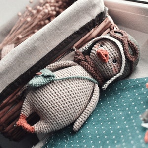 Pode incluir: Um brinquedo de pel&uacute;cia de pinguim de croch&ecirc; usando um chap&eacute;u de aviador marrom e &oacute;culos. O pinguim est&aacute; deitado em um tecido turquesa com bolinhas brancas.