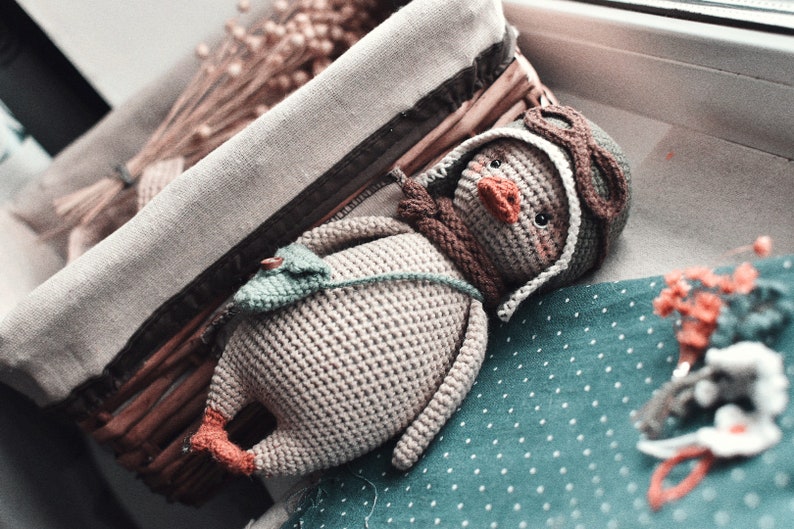 Pode incluir: Um brinquedo de pel&uacute;cia de pinguim de croch&ecirc; usando um chap&eacute;u de aviador marrom e &oacute;culos. O pinguim est&aacute; deitado em um tecido turquesa com bolinhas brancas.