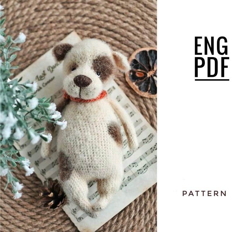 May include: Crochet pattern for a white and brown dog amigurumi toy. The pattern is available in English and is a PDF download. The toy is lying on a sheet of music.