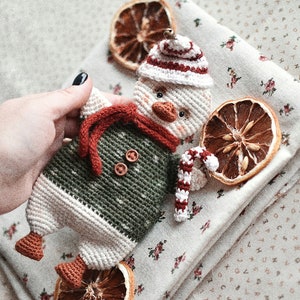 May include: A crocheted snowman toy with a red and white hat, green and white sweater, and a candy cane. The snowman is sitting on a white fabric with a floral pattern and dried orange slices.