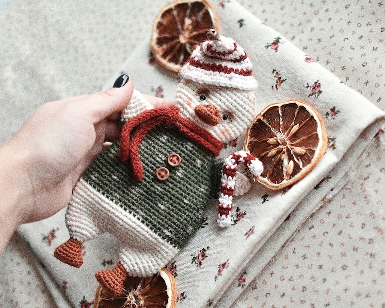 May include: A crocheted snowman toy with a red and white hat, green and white sweater, and a candy cane. The snowman is sitting on a white fabric with a floral pattern and dried orange slices.