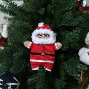 May include: A crocheted Santa Claus ornament with a red and white suit, black belt, and white beard. The ornament is hanging on a green Christmas tree.