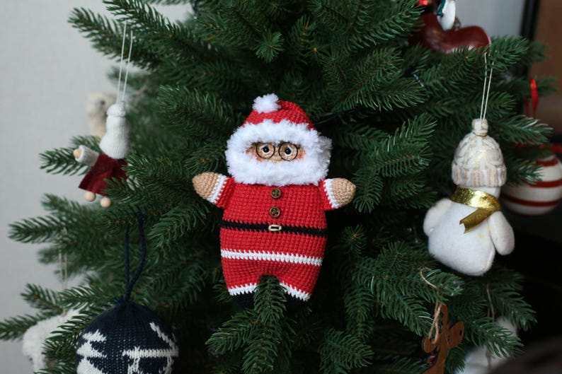 May include: A crocheted Santa Claus ornament with a red and white suit, black belt, and white beard. The ornament is hanging on a green Christmas tree.
