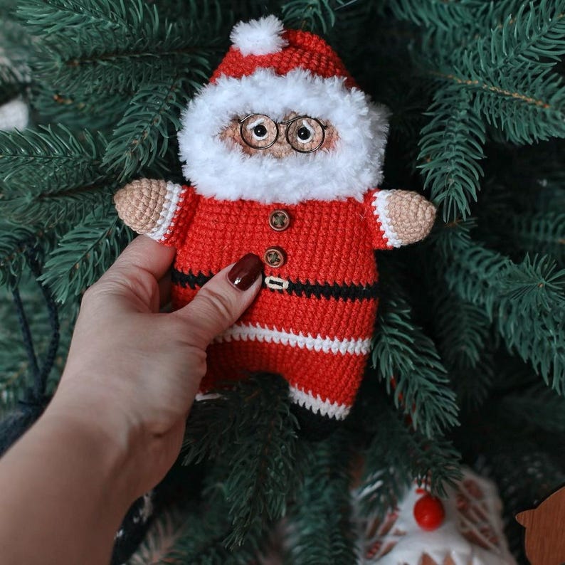 May include: A crocheted Santa Claus doll wearing a red suit with white trim, a black belt, and a white beard. The doll has brown eyes and a button nose.