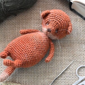 Op de afbeelding: Een handgemaakt, gehaakt oranje kattenknuffel met witte snorharen, liggend op een jute oppervlak. De kat heeft een lichtbruine neus en een gestreepte staart. Een bol groen garen, een naald en een schaar zijn ook zichtbaar.