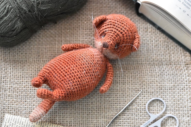 Op de afbeelding: Een handgemaakt, gehaakt oranje kattenknuffel met witte snorharen, liggend op een jute oppervlak. De kat heeft een lichtbruine neus en een gestreepte staart. Een bol groen garen, een naald en een schaar zijn ook zichtbaar.