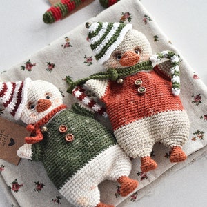 May include: Two crocheted penguin ornaments with red and white striped hats, one in green and white and the other in orange and white, with candy cane accents.  The text "made with love" is on a brown tag.