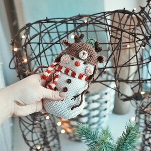 May include: A crocheted snowman wearing a brown reindeer hat with antlers and a red and white striped scarf. The snowman is holding a small brown stick in its hand.
