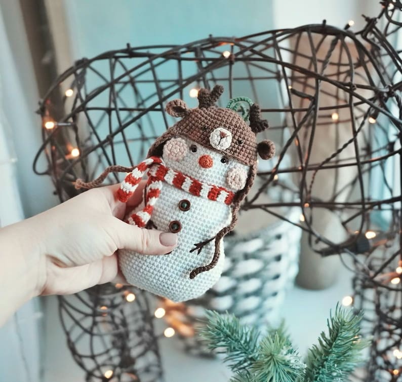 May include: A crocheted snowman wearing a brown reindeer hat with antlers and a red and white striped scarf. The snowman is holding a small brown stick in its hand.
