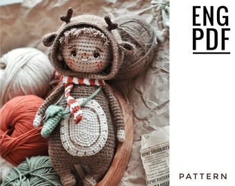 Może przedstawiać: Wzór na amigurumi na szydełku dla brązowego jelenia z czerwono-białą szarfą, zielonymi liśćmi i białym kółkiem na piersi. Wzór jest dostępny w języku angielskim i jest dostępny jako plik PDF do pobrania.