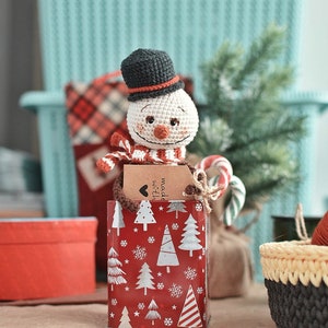 Puede incluir: Un mu&ntilde;eco de nieve de crochet con un sombrero negro y una bufanda roja y blanca est&aacute; sentado en una caja de regalo roja y blanca con una etiqueta que dice "hecho con amor".