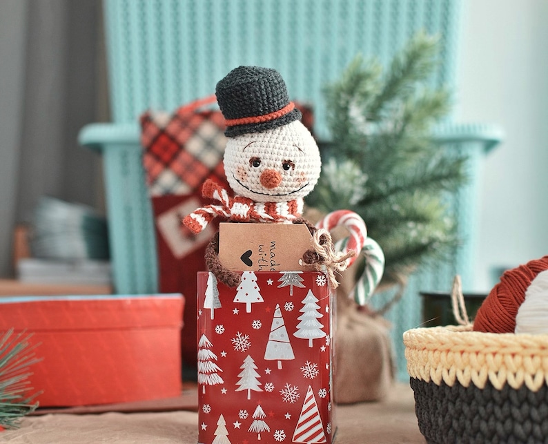 Puede incluir: Un mu&ntilde;eco de nieve de crochet con un sombrero negro y una bufanda roja y blanca est&aacute; sentado en una caja de regalo roja y blanca con una etiqueta que dice "hecho con amor".