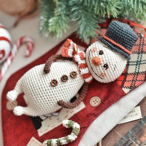 Puede incluir: Un mu&ntilde;eco de nieve de crochet con un sombrero gris, una bufanda a rayas rojas y blancas y botones marrones. El mu&ntilde;eco de nieve est&aacute; tumbado sobre una tela de cuadros rojos y negros.