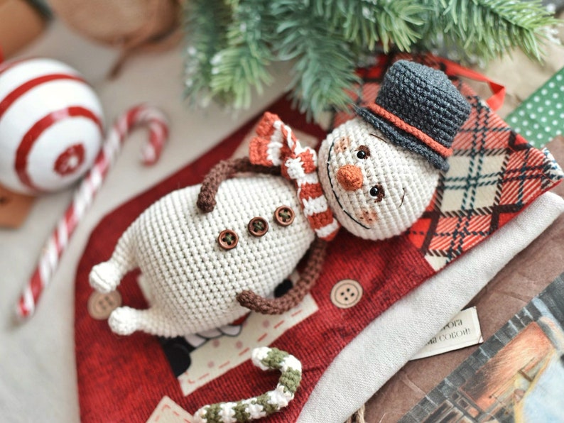 Puede incluir: Un mu&ntilde;eco de nieve de crochet con un sombrero gris, una bufanda a rayas rojas y blancas y botones marrones. El mu&ntilde;eco de nieve est&aacute; tumbado sobre una tela de cuadros rojos y negros.