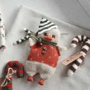 May include: A crocheted duck wearing a green and white striped hat and a red and white striped scarf. The duck is holding a green and white candy cane in its beak. There are two other candy canes in the image, one brown and white and one red and green.