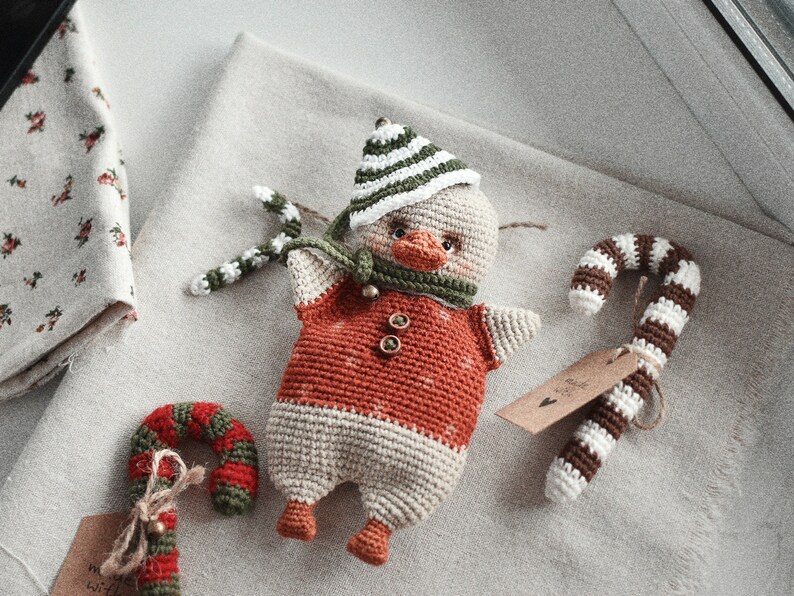 May include: A crocheted duck wearing a green and white striped hat and a red and white striped scarf. The duck is holding a green and white candy cane in its beak. There are two other candy canes in the image, one brown and white and one red and green.