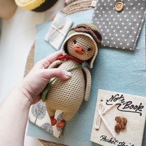 Pode incluir: Um brinquedo de peluche de croch&ecirc; em forma de pato que usa um chap&eacute;u de aviador verde e &oacute;culos de prote&ccedil;&atilde;o. O pato est&aacute; segurando uma al&ccedil;a verde em sua asa direita. O pato &eacute; bege com p&eacute;s laranja.