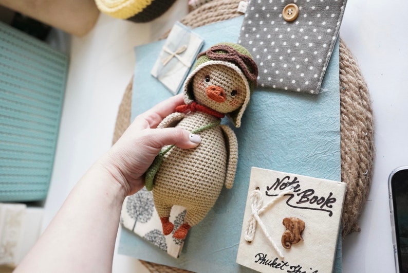 Pode incluir: Um brinquedo de peluche de croch&ecirc; em forma de pato que usa um chap&eacute;u de aviador verde e &oacute;culos de prote&ccedil;&atilde;o. O pato est&aacute; segurando uma al&ccedil;a verde em sua asa direita. O pato &eacute; bege com p&eacute;s laranja.