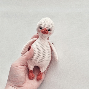 Pode incluir: Um brinquedo de pato de croch&ecirc; branco com p&eacute;s vermelhos e olhos pretos. O pato est&aacute; sendo segurado em uma m&atilde;o.