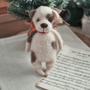 May include: A crocheted white and brown dog toy with a red scarf. The toy is standing on a sheet of music with the text "Just One Last Dance" printed on it.