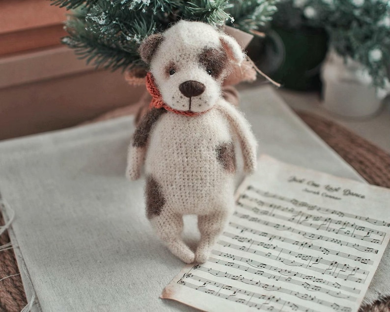 May include: A crocheted white and brown dog toy with a red scarf. The toy is standing on a sheet of music with the text "Just One Last Dance" printed on it.