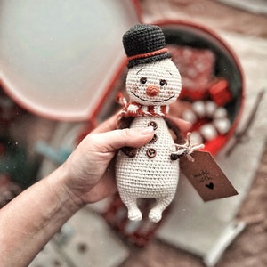Puede incluir: Un juguete de mu&ntilde;eco de nieve de crochet con un sombrero de copa negro, una bufanda roja y botones marrones. El mu&ntilde;eco de nieve est&aacute; siendo sostenido por una mano y tiene una etiqueta que dice "hecho con amor".
