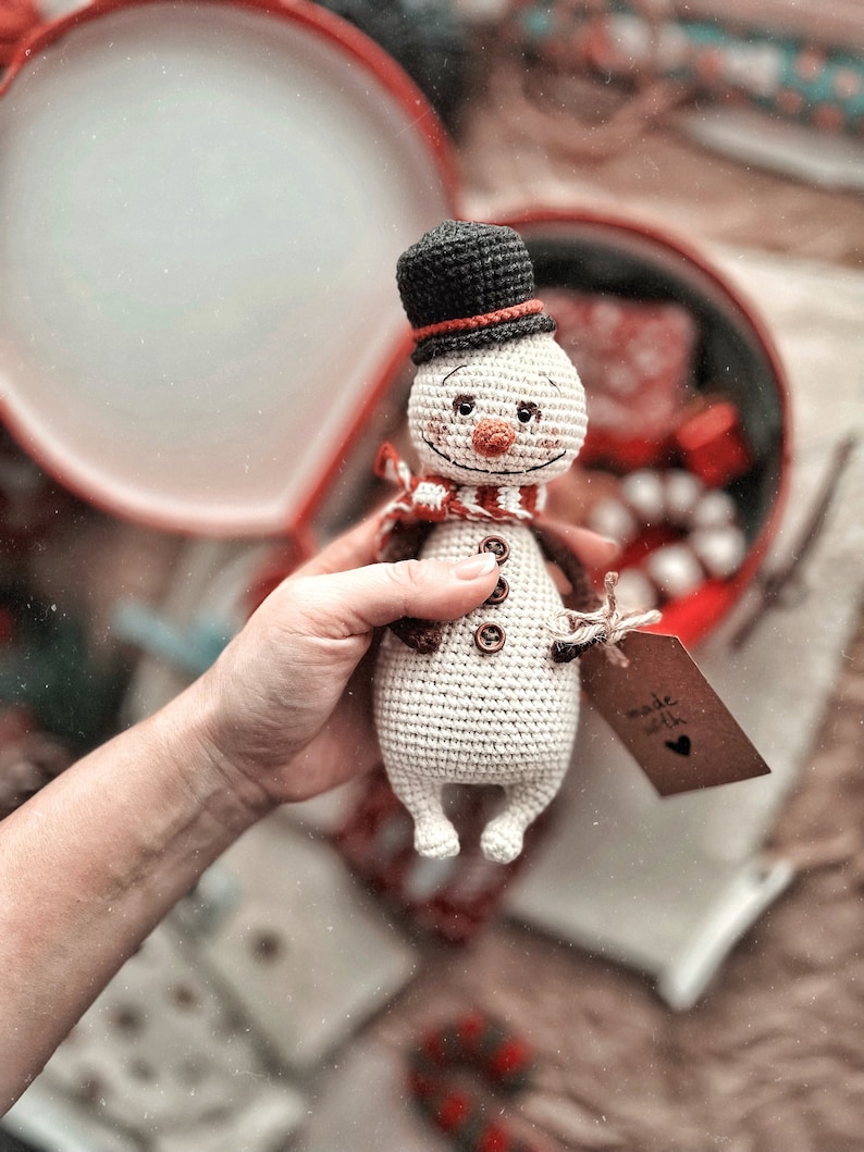Puede incluir: Un juguete de mu&ntilde;eco de nieve de crochet con un sombrero de copa negro, una bufanda roja y botones marrones. El mu&ntilde;eco de nieve est&aacute; siendo sostenido por una mano y tiene una etiqueta que dice "hecho con amor".
