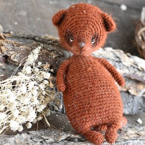 Op de afbeelding: Een handgemaakte bruine gehaakte teddybeerpop met kleine oren, een puntneus en donkere ogen. De beer staat op een stuk hout met gedroogde witte bloemen en een takkenkrans op de achtergrond. Een uniek, handgemaakt item.