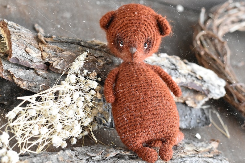 Op de afbeelding: Een handgemaakte bruine gehaakte teddybeerpop met kleine oren, een puntneus en donkere ogen. De beer staat op een stuk hout met gedroogde witte bloemen en een takkenkrans op de achtergrond. Een uniek, handgemaakt item.