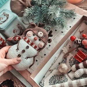 May include: A crocheted snowman ornament with a brown reindeer hat and a red and white striped scarf. The ornament is sitting in a box with a white and brown patterned fabric.