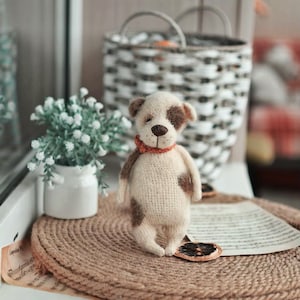 May include: A crocheted dog toy with brown spots, wearing a red collar, sitting on a brown woven mat. The toy is on a windowsill with a white pot of small white flowers and a wicker basket in the background.