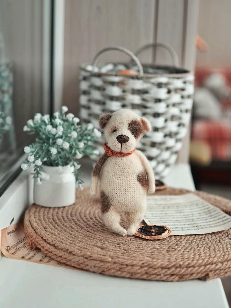May include: A crocheted dog toy with brown spots, wearing a red collar, sitting on a brown woven mat. The toy is on a windowsill with a white pot of small white flowers and a wicker basket in the background.