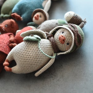 Pode incluir: Um pinguim de brinquedo de croch&ecirc; usando um chap&eacute;u verde e marrom com uma borda branca. O pinguim tem um bico marrom e olhos pretos. O pinguim est&aacute; deitado em uma superf&iacute;cie cinza.