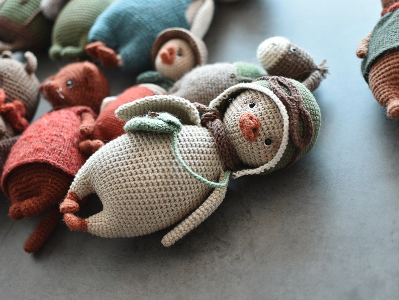 Pode incluir: Um pinguim de brinquedo de croch&ecirc; usando um chap&eacute;u verde e marrom com uma borda branca. O pinguim tem um bico marrom e olhos pretos. O pinguim est&aacute; deitado em uma superf&iacute;cie cinza.
