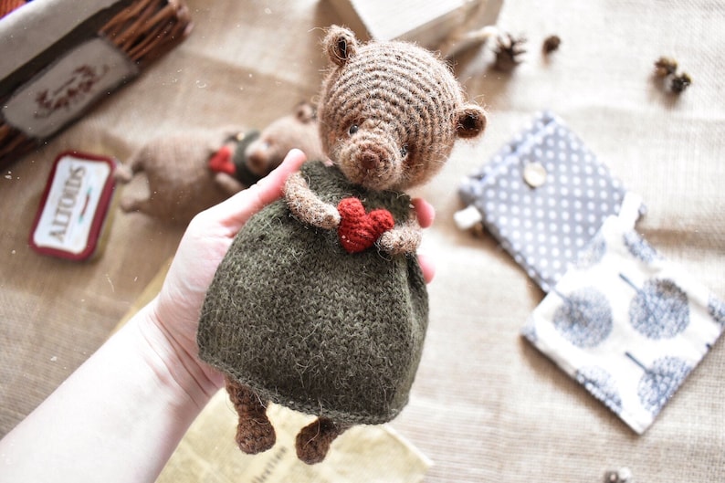 May include: A brown crocheted teddy bear wearing a green dress and holding a red heart.