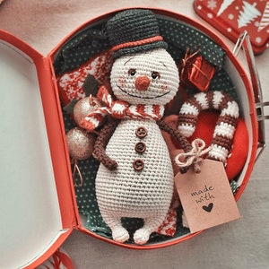 Puede incluir: Un adorno de mu&ntilde;eco de nieve de crochet en una caja de regalo roja y blanca con un bast&oacute;n de caramelo y una etiqueta que dice "hecho con amor".