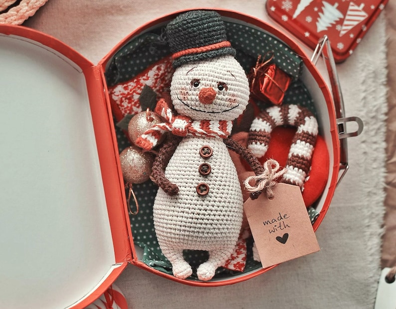 Puede incluir: Un adorno de mu&ntilde;eco de nieve de crochet en una caja de regalo roja y blanca con un bast&oacute;n de caramelo y una etiqueta que dice "hecho con amor".
