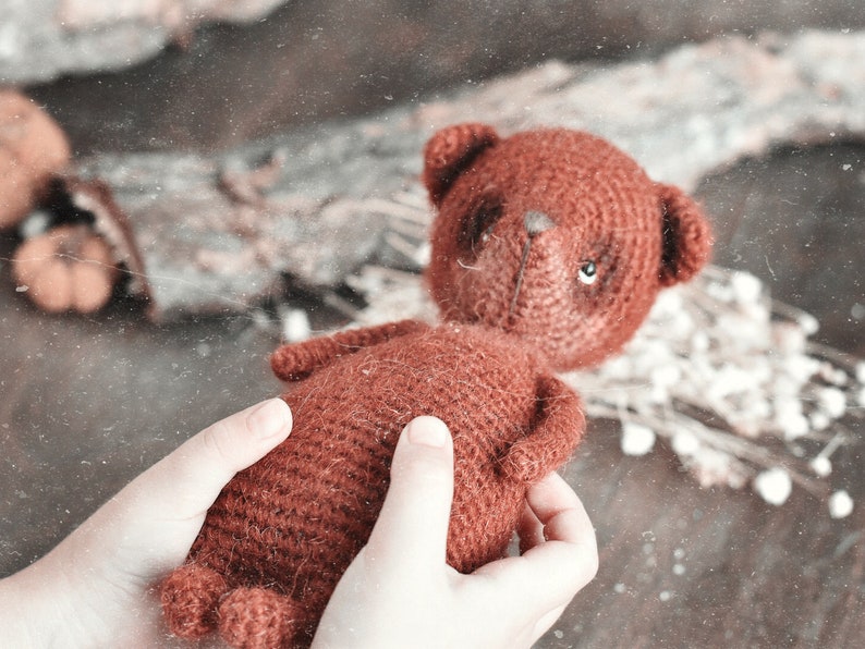 Op de afbeelding: Een handgemaakte, roodbruine gehaakte teddybeer met kleine, ronde oren en een gestikte neus. De beer wordt in de handen van een persoon gehouden, met een vage achtergrond van gedroogde bloemen en bladeren.