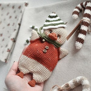 May include: A crocheted toy duck wearing a green and white hat and a red and white sweater. The duck is holding a brown and white crocheted candy cane.