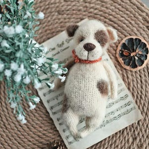May include: A crocheted white and brown dog toy with a red collar. The toy is lying on a sheet of music with a dried orange slice on a woven surface.