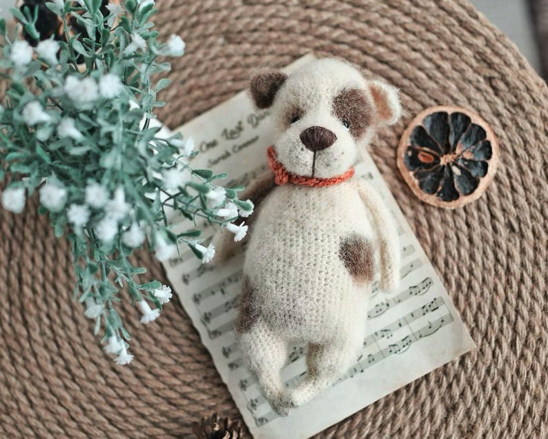 May include: A crocheted white and brown dog toy with a red collar. The toy is lying on a sheet of music with a dried orange slice on a woven surface.