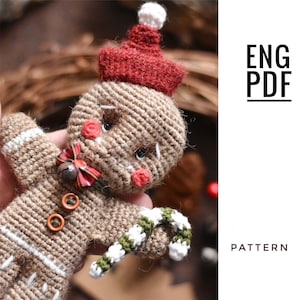 Puede incluir: Adorno de jengibre de crochet con un gorro rojo, un bastón de caramelo blanco y verde y una pajarita marrón. El adorno es marrón con rayas blancas y tiene una cara cosida con mejillas rojas. ENG PDF PATTERN.