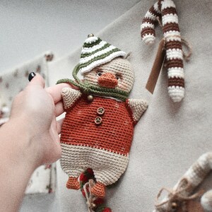 May include: A crocheted stuffed toy duck wearing a green and white striped hat and a red and white sweater. The duck is holding a candy cane in its beak.