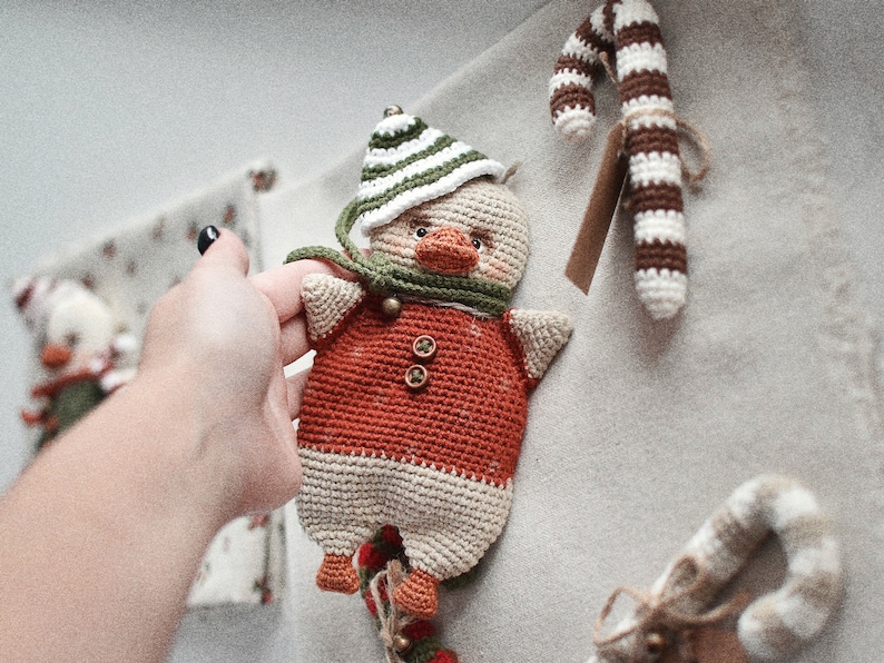 May include: A crocheted stuffed toy duck wearing a green and white striped hat and a red and white sweater. The duck is holding a candy cane in its beak.