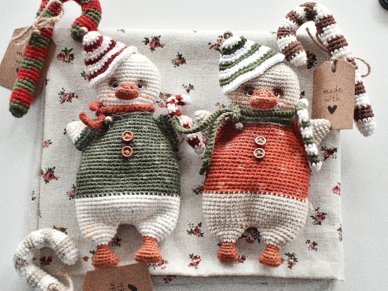 May include: Two crocheted snowman ornaments with candy cane accents. The snowman on the left is wearing a green and white outfit, while the snowman on the right is wearing a red and white outfit. Both snowmen have black eyes and a black button nose.