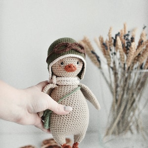 Pode incluir: Um pato de brinquedo de croch&ecirc; vestindo um chap&eacute;u de aviador verde com &oacute;culos marrons e um cachecol branco. O pato est&aacute; segurando uma bolsa de croch&ecirc; verde.