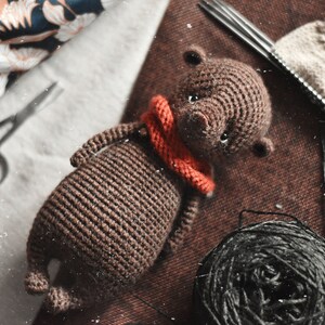 May include: A brown crocheted bear with a red scarf. The bear is lying on a brown fabric surface with a ball of black yarn and knitting needles.