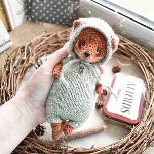 Op de afbeelding: Een handgemaakte gehaakte vossenpop met een lichtgroene jumpsuit met capuchon. De vos heeft bruine vacht en een zwarte neus. De pop wordt vastgehouden in de buurt van een takkenkrans en een klein Altoids-blikje.