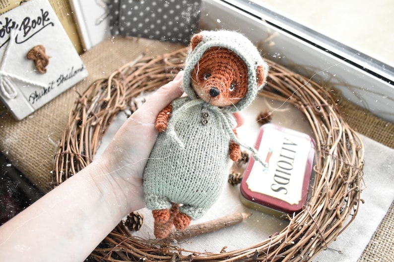 Op de afbeelding: Een handgemaakte gehaakte vossenpop met een lichtgroene jumpsuit met capuchon. De vos heeft bruine vacht en een zwarte neus. De pop wordt vastgehouden in de buurt van een takkenkrans en een klein Altoids-blikje.